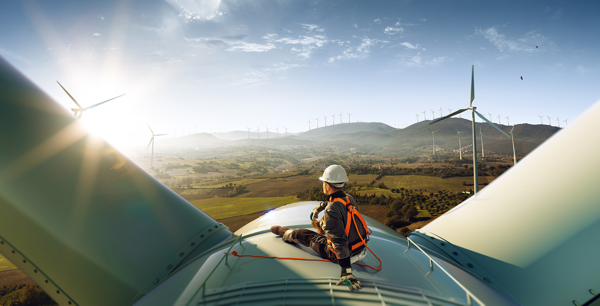 An engineer sits outside on a wind turbine in the sun looking across green fields towards a wind farm