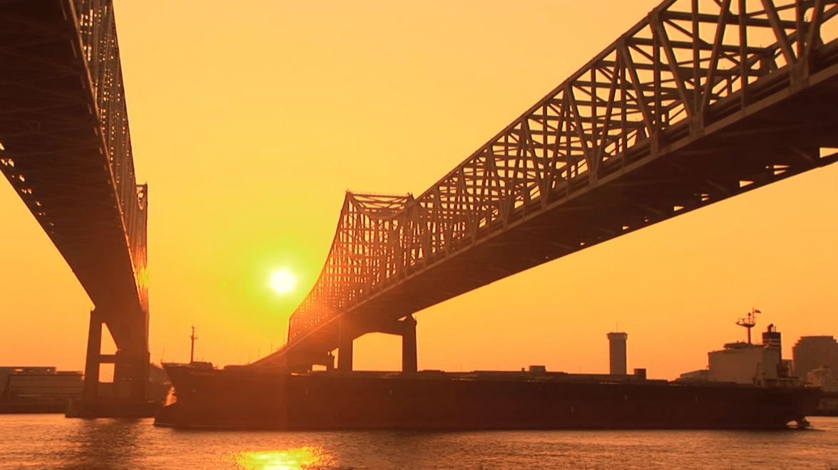 Silhouetted construction workers on a steel framework against an orange sunset sky.